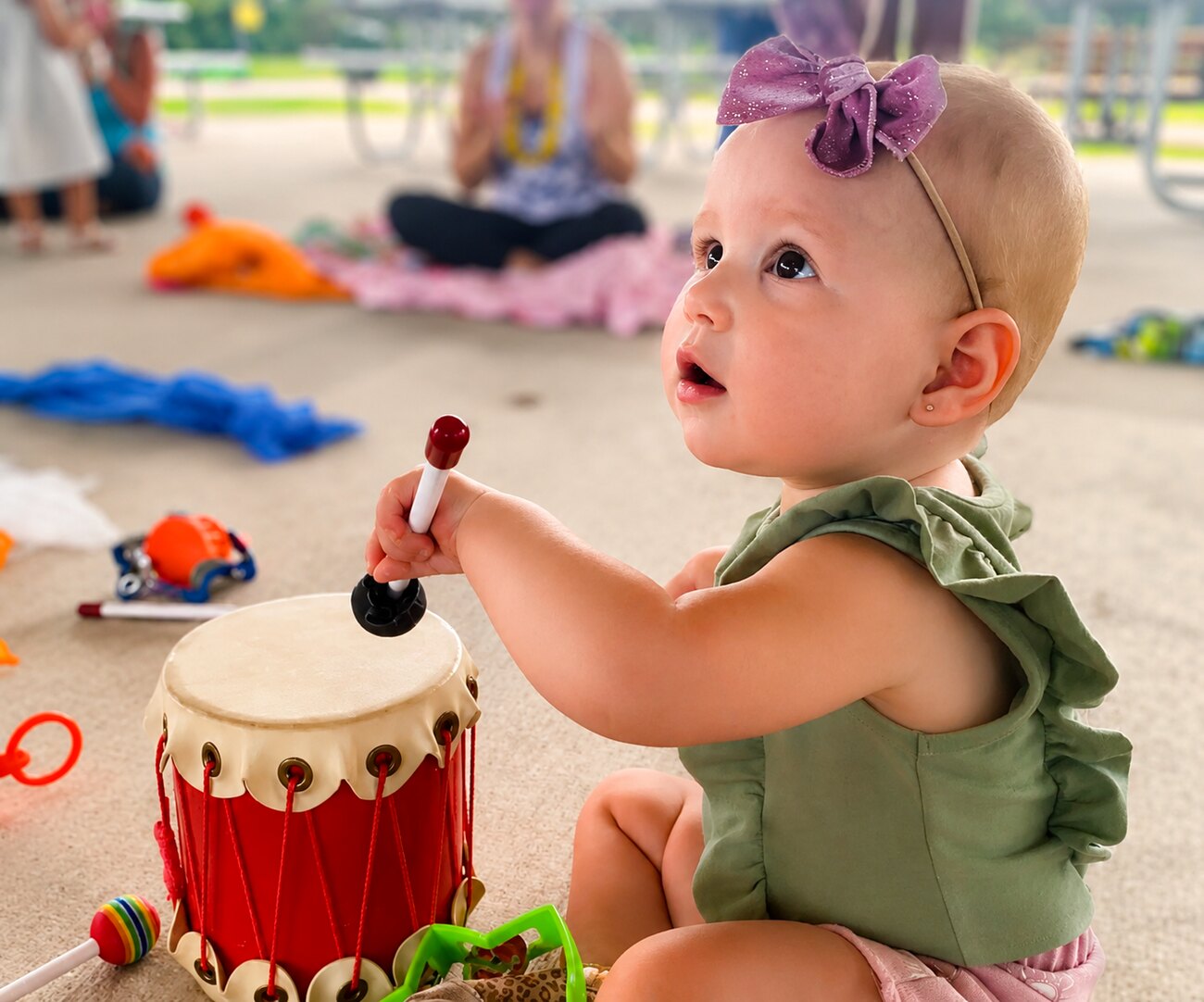 A baby joyfully playing a small red hand drum at a Songbirds Music class in Liberty Station, San Diego